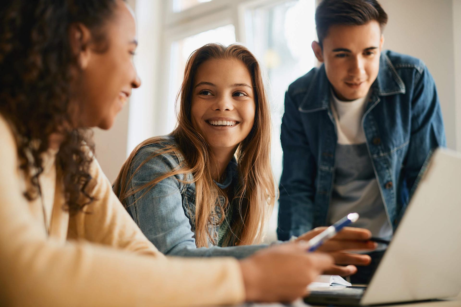 Three young adults collaborate cheerfully, gathered around a laptop. A woman with a pen smiles at her friends, set in a brightly lit room.