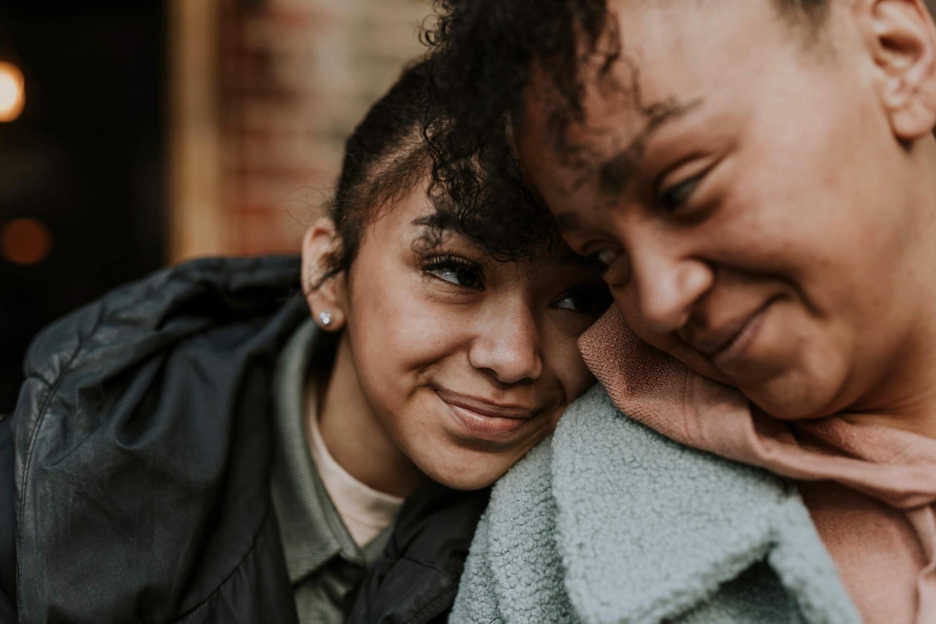 Close-up of two people sharing a tender moment; one leans on the other's shoulder, both smiling warmly. Cozy jackets add a sense of comfort and bonding.