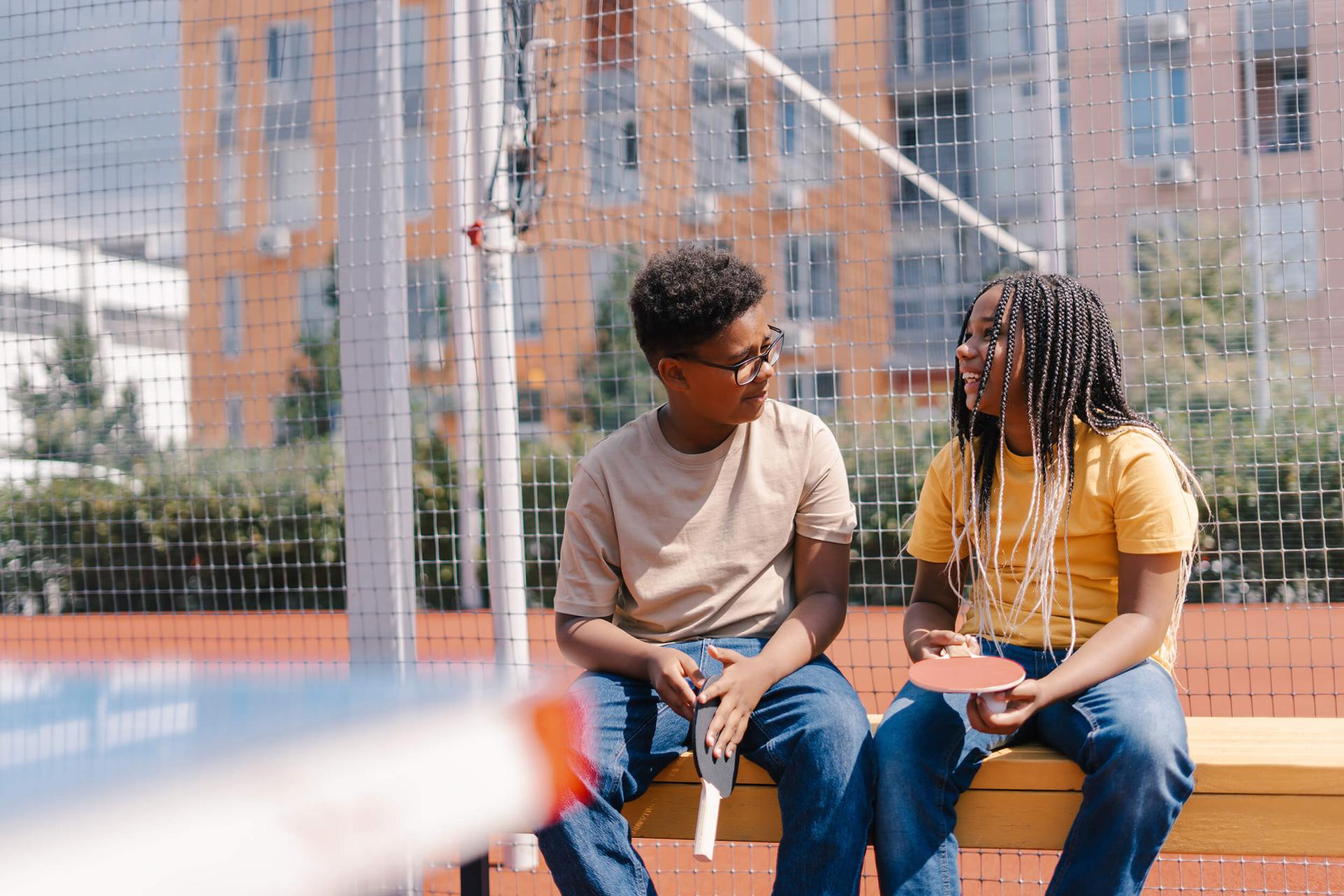 Two kids sit on a bench at a playground, holding ping pong paddles, smiling at each other. Urban buildings and a netted sport court are in the background.