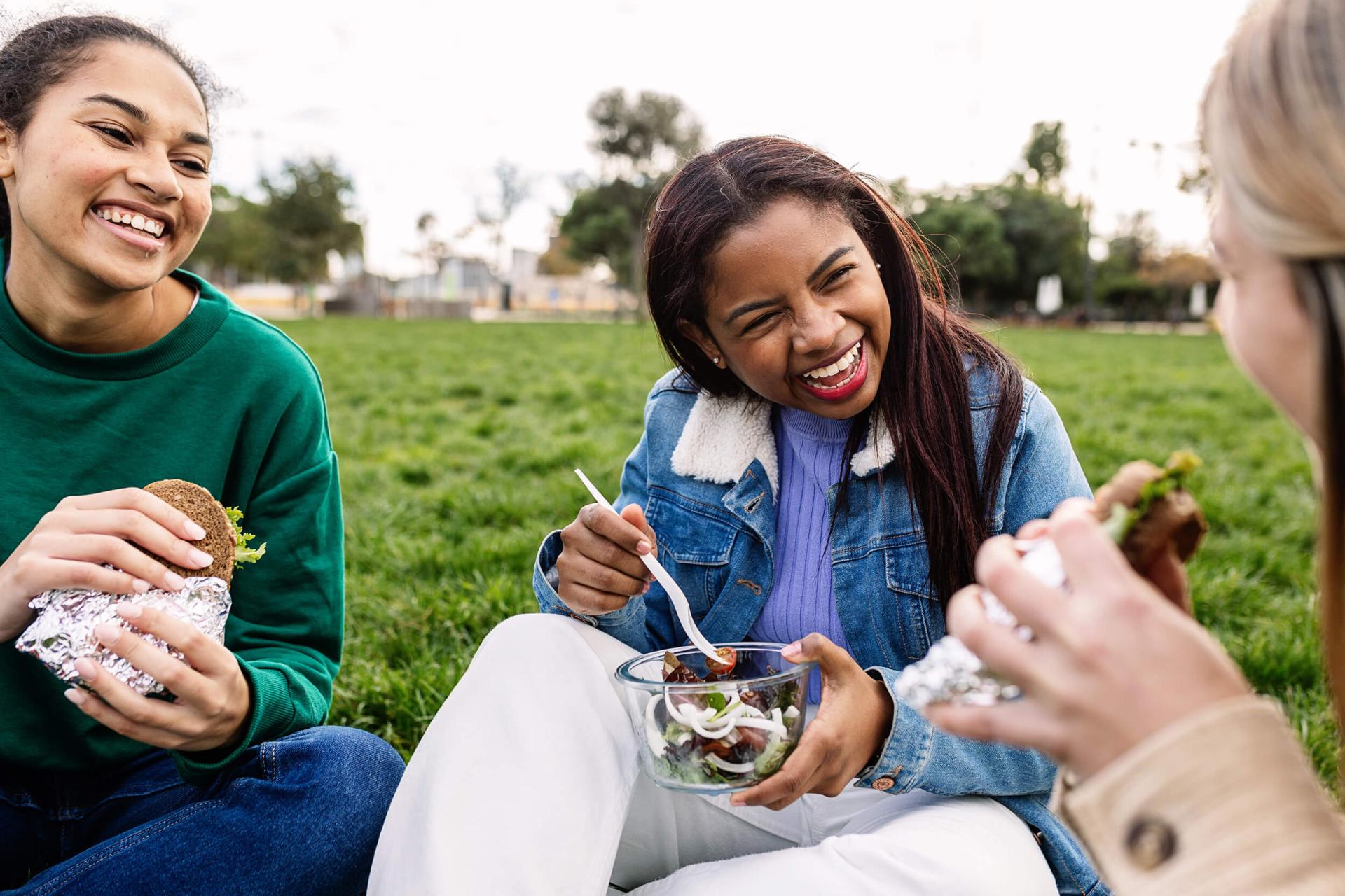 Three friends enjoy a picnic on the grass, smiling and laughing. One holds a sandwich, another a salad, conveying joy and relaxation in the park.