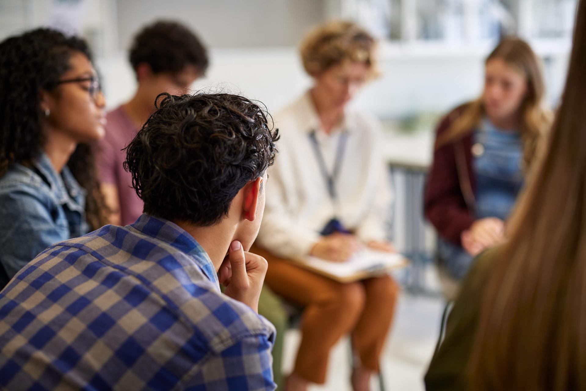 A diverse group of people sits in a circle, engaged in discussion. The setting is informal and collaborative, with a focus on listening and sharing.