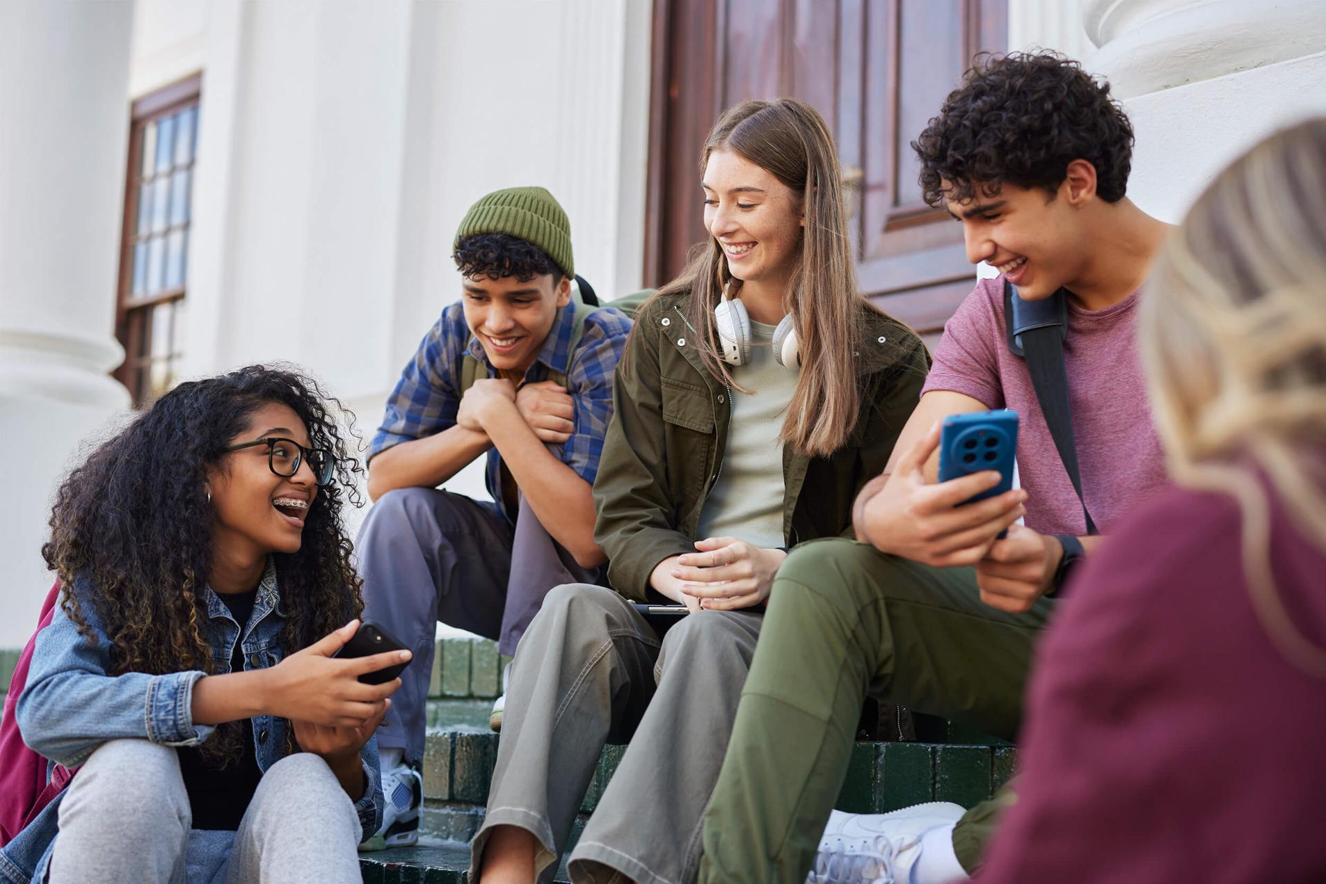 A group of four diverse, smiling teenagers sit on outdoor steps, chatting and holding smartphones, conveying a sense of friendship and connection.