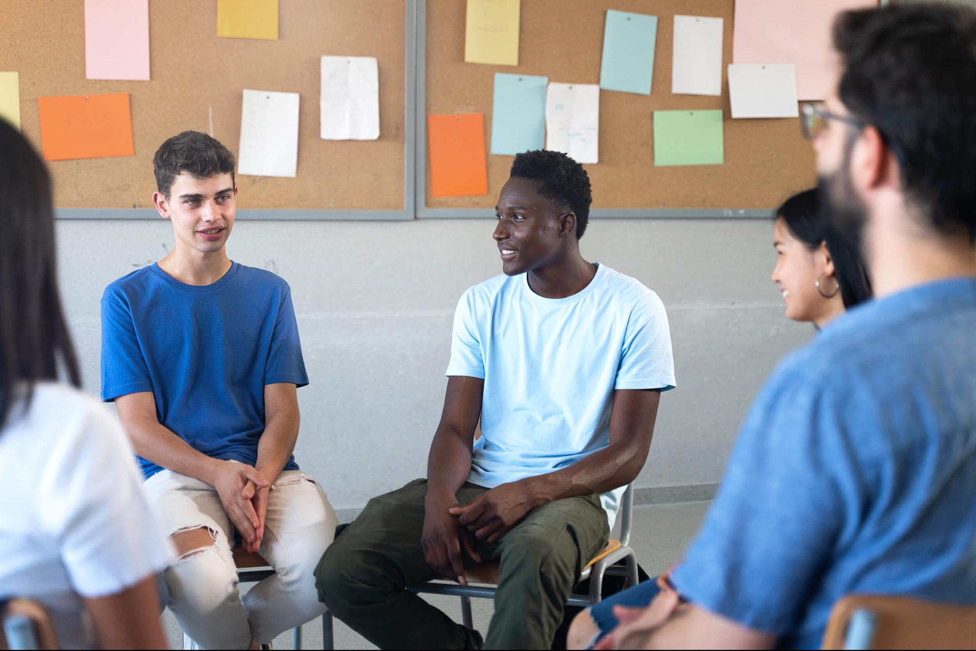 A group of diverse young adults sit in a circle, engaging in a lively discussion. The room has a bulletin board with colorful papers, creating a warm, collaborative atmosphere.