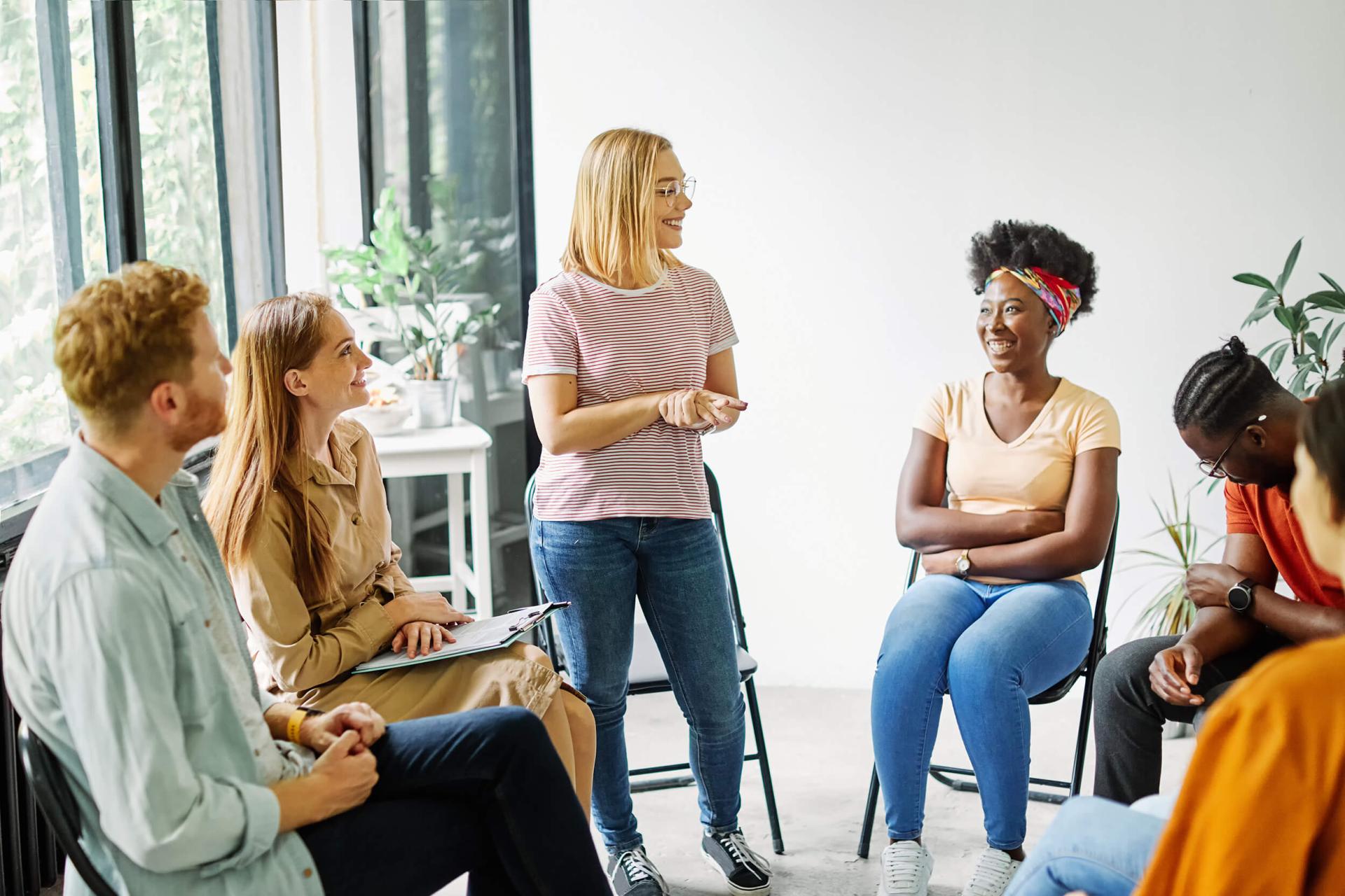 A diverse group of six people in a casual meeting room sit in a circle, engaged in discussion. A woman stands, smiling, conveying a positive, collaborative mood.