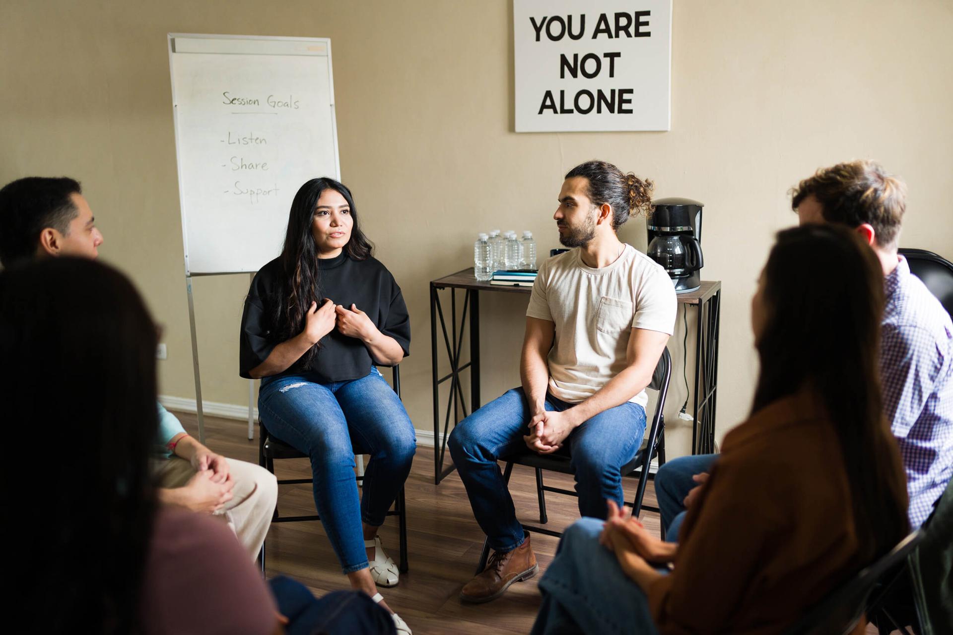 A diverse group sits in a supportive circle at a meeting. A woman speaks, expressing herself. A sign reads 