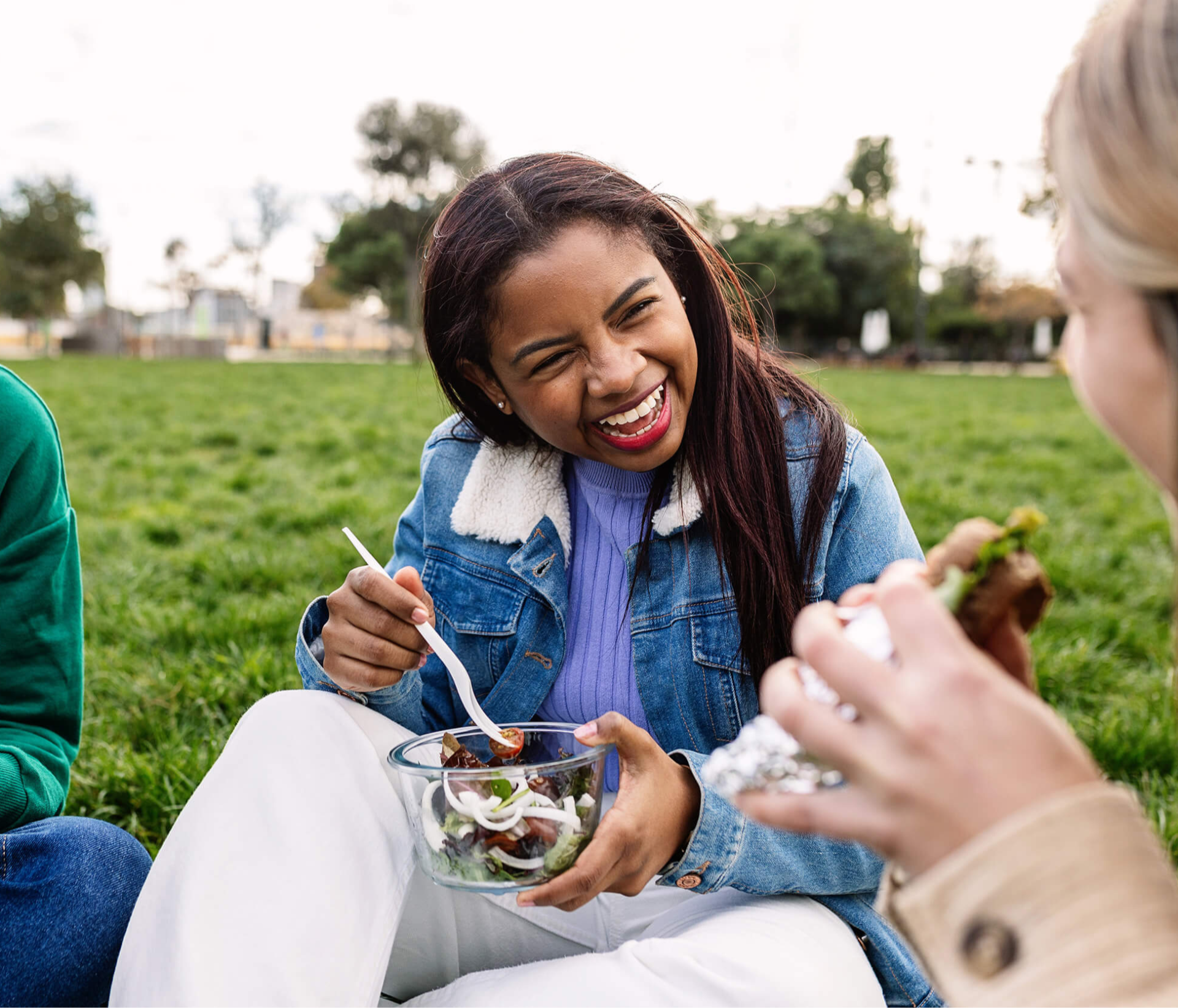 Three friends enjoy a picnic on the grass, smiling and laughing. One holds a sandwich, another a salad, conveying joy and relaxation in the park.
