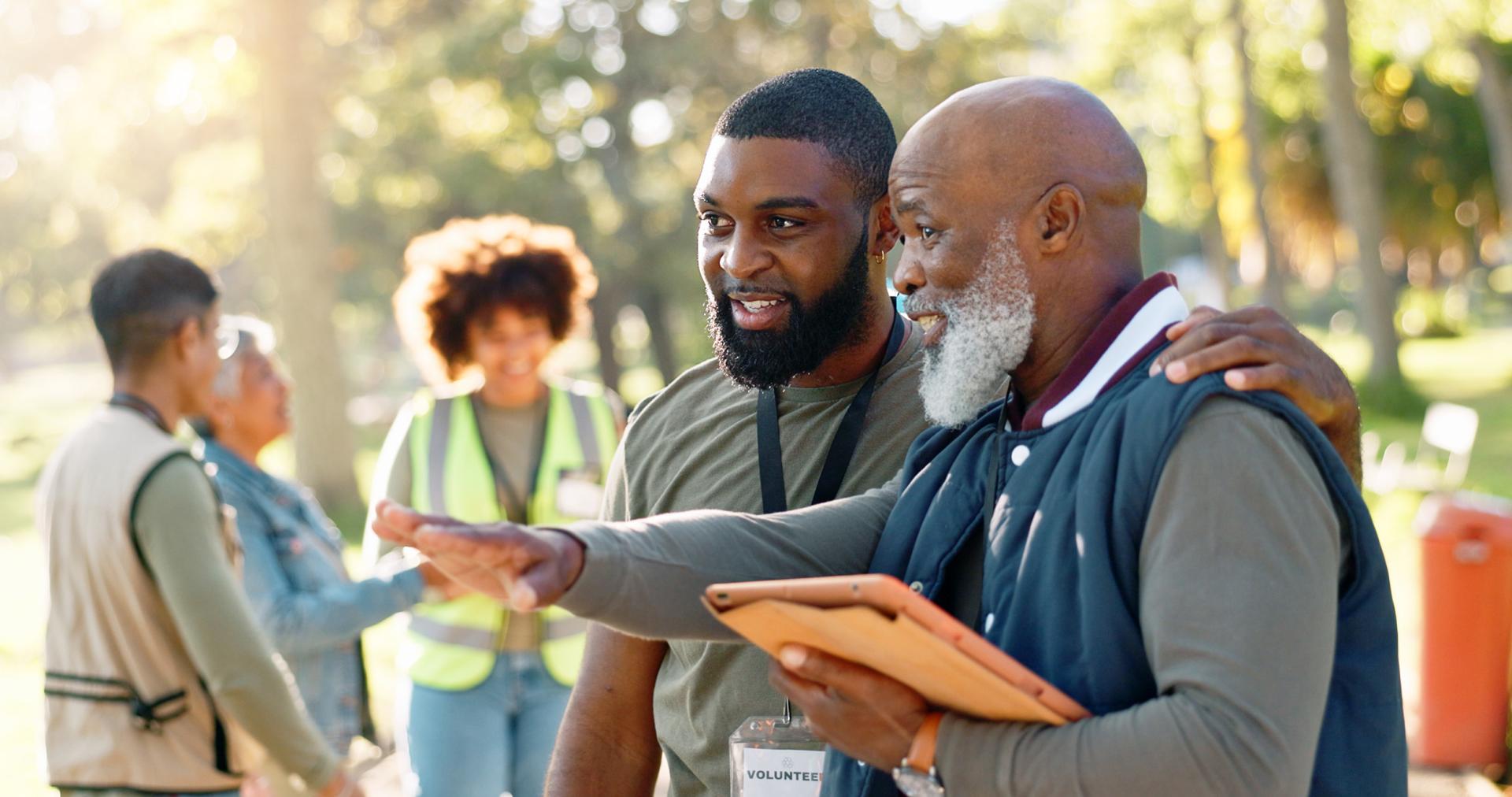 Two men, smiling, discuss something outdoors in a sunlit park. One gestures ahead while holding a tablet. Others in the background wear safety vests.