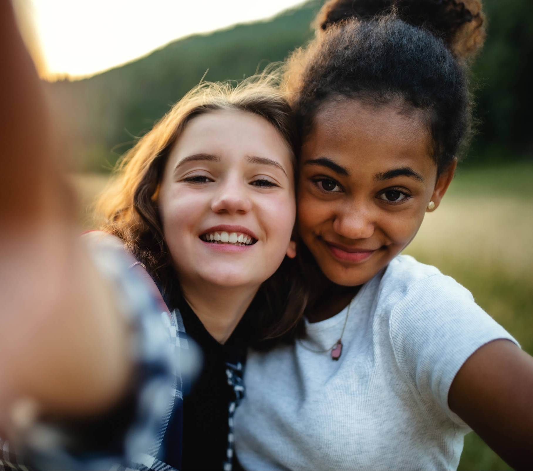 Two smiling young women taking a selfie in a scenic outdoor setting. The mood is joyful and carefree, with greenery blurred in the background.