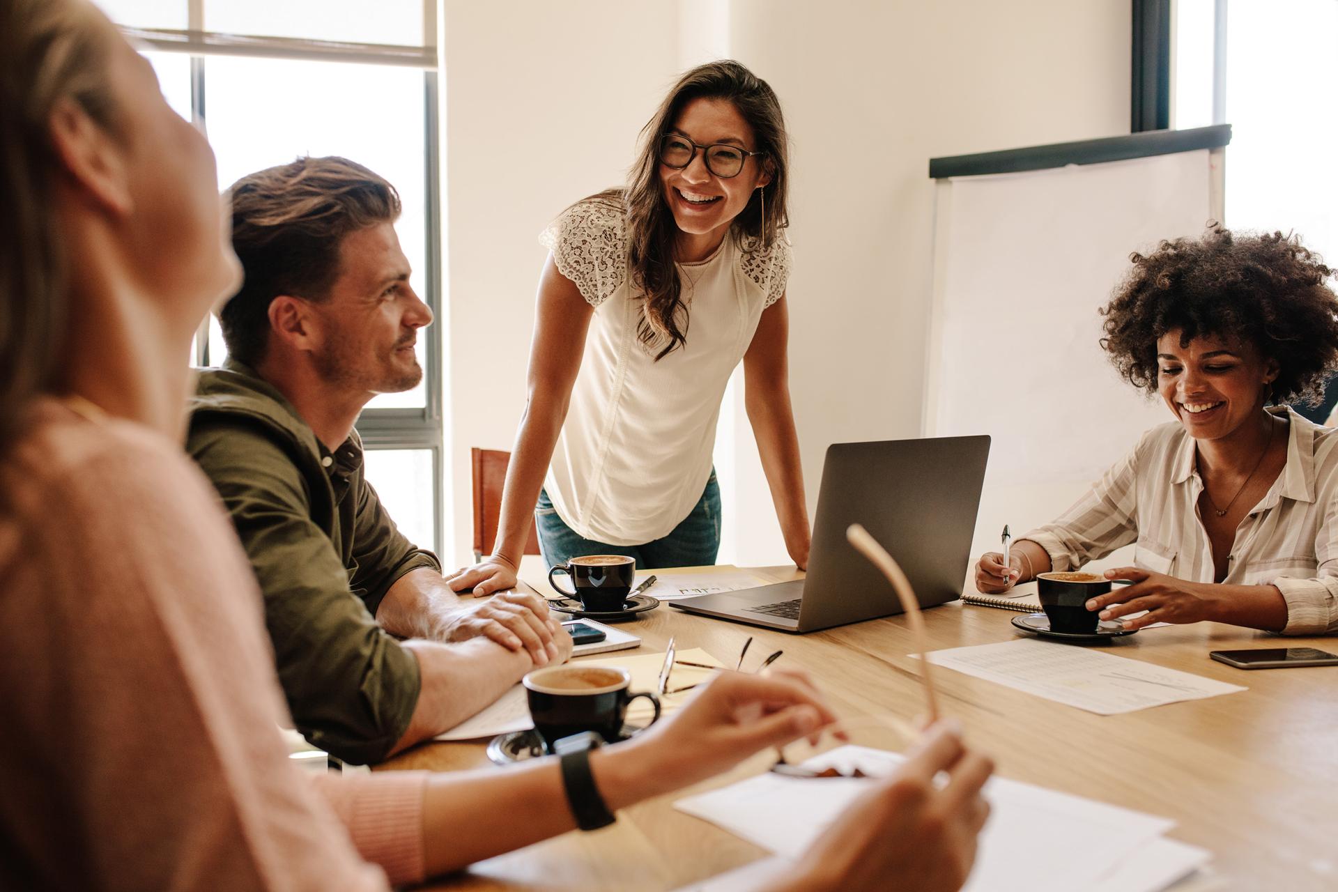 A diverse group of four people engaged in a lively meeting around a table. They are smiling, with coffee cups, papers, and a laptop present, conveying a collaborative and positive atmosphere.